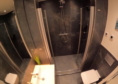Das Badezimmer mit Steinfliesen, Waschbecken, Toilette, Spiegelschrank, ebenerdige Dusche mit großem Duschkopf, Fußbodenheizung,