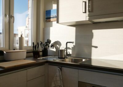 Blick in die Küche, mit Spüle, Messern, Holzbrett, viel Licht durch das Fenster, Schränke, kleines Bild an der Wand