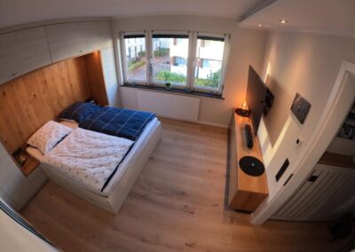 Schlafzimmer mit großem Bett für zwei Personen, Bettdecken, Kissen, Holzfußboden, Blickrichtung zum Fenster zur Straße, Wandschränke, gedimmtes Licht,