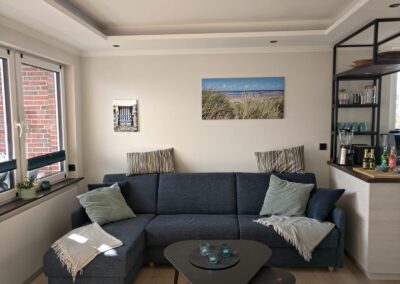 gemütliche Couch im Wohnzimmer, viele Kissen, Wolldecken auf der Couch und im Reclaim, Fenster an der Seite, offene Küche an der anderen Seite